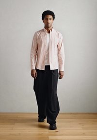 Ανοιχτό ροζ πουκάμισο με κουμπιά, φορεμένο πάνω από λευκό T-shirt, συνδυασμένο με φαρδιά μαύρη παντελόνια και μαύρα παπούτσια, πάνω σε απλό φόντο.