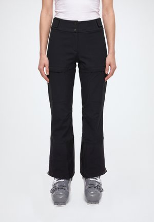 Modèle portant un pantalon de ski noir évasé avec des poches sur les cuisses et des chaussures de ski grises, sur un fond blanc uni.