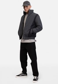 Giubbotto piumino nero e grigio con colletto alto, tessuto testurizzato, tasche laterali, abbinato a una maglietta grigia, pantaloni neri e sneaker nere.