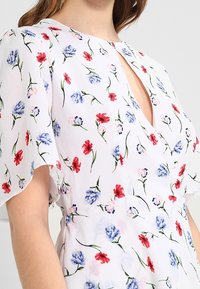 Vit klänning med små blommönster i röd, blå och rosa färg, med fladderärmar och nyckelhalsringning, bärs av en person.