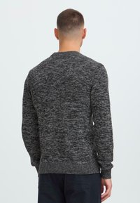 Man met kort haar, gekleed in een donkergrijze gemarmerde trui van gebreid materiaal en zwarte broek, kijkt weg tegen een zachte lichte achtergrond.