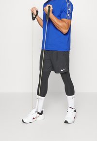 Modrá košile s krátkým rukávem, černé šortky, černé legíny, bílé ponožky a bílé tenisky s červenými akcenty. Dvě odporové gumy drženy v rukou.