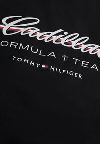 Λογότυπο Cadillac με κόκκινη, λευκή και μπλε λωρίδα πάνω από το κείμενο "Formula 1 Team" και "Tommy Hilfiger" με λογότυπο σημαίας σε μαύρο ύφασμα.