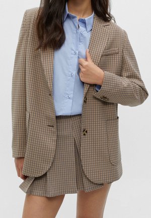 Femme portant un blazer et une jupe à carreaux beige et marron avec une chemise bleu clair boutonnée, ajustant le blazer d'une main.