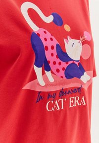 Czerwona koszulka z grafiką rozciągającego się kota w niebieskim i różowym kolorze w kropki, z napisem "W mojej erze kota w dół."