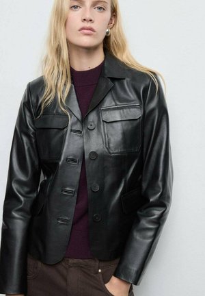 Blonde Frau, die eine schwarze Lederjacke mit Knöpfen über einem dunkelpurpurnen Pullover und braunen Hosen trägt, vor einem hellen Hintergrund steht.