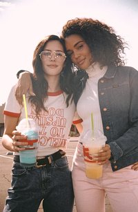 Δύο γυναίκες στέκονται κοντά η μία στην άλλη, κρατώντας η καθεμία ένα μεγάλο ποτό. Η μία φοράει ένα λευκό t-shirt με κόκκινες λεπτομέρειες και τζιν ψηλής μέσης. Η άλλη έχει έναν λαιμόκοψη και ένα τζιν μπουφάν.