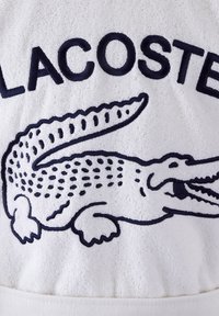Tissu en éponge blanche avec un logo crocodile brodé en bleu marine et l'inscription "LACOSTE" au-dessus, avec des textures et des accents détaillés.