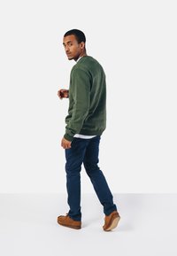 Grøn sweatshirt med rund hals, parret med navy blå bukser og brune sko. Modellen står let drejet til venstre.