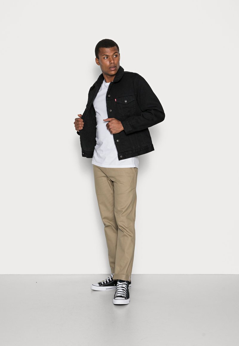 Czarna kurtka dżinsowa z podszyciem z sherpy, z zapięciem na zatrzaski, czterema kieszeniami, noszona na białej koszulce, w połączeniu z khaki spodniami i czarnymi trampkami.