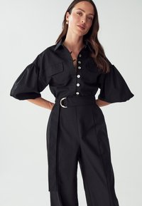Chemise noire à col avec des manches bouffantes, des poches sur le devant et une fermeture à boutons. Stylisée avec un pantalon noir taille haute et une boucle de ceinture circulaire.