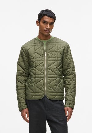 Mand iført en olivengrøn quiltet jakke med lynlås og sorte bukser, stående foran en ensartet hvid baggrund.