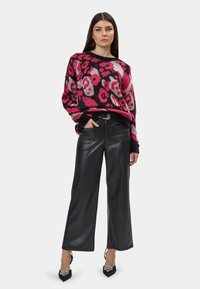 Flauschiger, übergroßer Pullover in Schwarz mit einem pinken und roten Blumenmuster. Schwarze, weite Hosen aus Kunstleder und spitze Absatzschuhe mit Verzierungen.