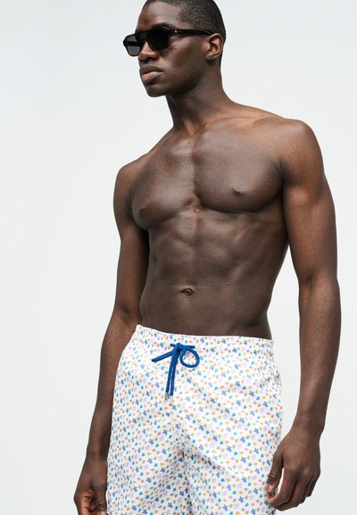 Μυώδης άνδρας χωρίς πουκάμισο, φορώντας μαύρα γυαλιά ηλίου και λευκά μαγιό με πολύχρωμο ανθισμένο σχέδιο και μπλε κορδόνι.