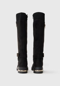 Schwarze Wildleder-Stiefel bis zum Knie mit einem gerippten Stoffoberteil, einem seitlichen Reißverschluss und einem metallischen Akzent an der Ferse, ausgestattet mit einem Riemen mit einer Schnalle.