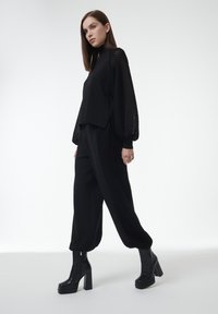 Vrouw in zwarte hoge hakken, losse zwarte broek met gestructureerde zijpanelen, en een zwarte top met mesh-gepatenteerde ballonsleeves, staand.