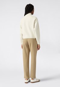 Giacca leggera color crema con colletto alto, abbinata a pantaloni beige. Le scarpe sono bianche con dettagli marroni e un logo. Materiali lisci in tutto l'outfit.
