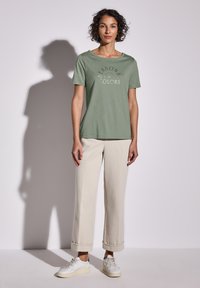 Zelené bavlněné tričko s potiskem "Sezónní barvy", kombinované se světle béžovými bavlněnými kalhotami a bílými teniskami. Jednoduchý design, volný střih.