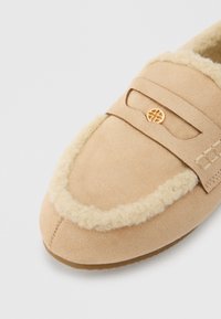 Μπεζ παντόφλα μοκασίνι με μαλακό σουέτ επάνω μέρος, αφράτη επένδυση από σιέρλινγκ, διακοσμητική ταινία με χρυσή εμβλήμα και ελαστική σόλα.