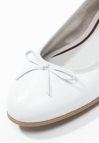 Ballerine en cuir blanc avec un embout rond, ornée d'un petit nœud à l'avant et d'une semelle en caoutchouc brun clair. Texture lisse.