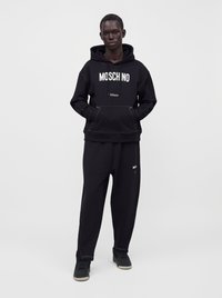 Homme debout, les mains dans les poches, portant un sweat à capuche Moschino noir et un pantalon noir assorti sur fond blanc uni.