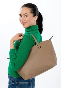 Béžová kožená kabelka s texturovaným povrchem, dvěma pevnými uchy a zipem na vrchu. Modelka má na sobě zelený žebrovaný rolák a pózuje z boku.