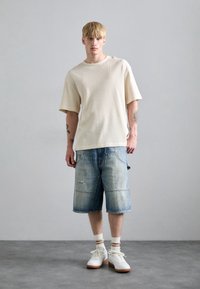 Chemise à manches courtes texturée beige clair, associée à un short en denim bleu clair avec des détails usés, portée avec des baskets blanches et des chaussettes rayées.