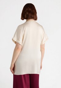 Blusa colore crema a maniche corte con vestibilità ampia; tessuto morbido e liscia; presenta maniche arrotolate e un orlo curvo.