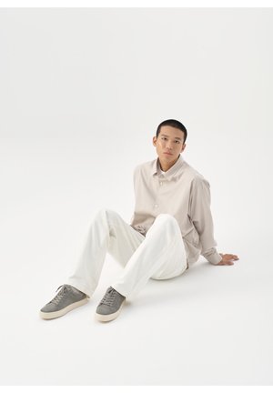 Ung mand sidder på gulvet iført en beige skjorte, hvide bukser og grå sneakers, kigger frem med et neutralt udtryk.