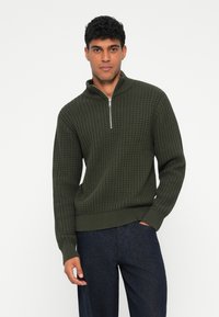 Pulover tricotat cu guler cu fermoar, de culoare verde închis. Prezintă un model texturat și manșete și tiv crantă. Poartă pantaloni închiși la culoare.