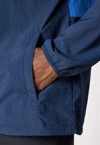 Veste légère bleu marine avec une poche latérale, un poignet élastique et une texture lisse. Présente des accents bleus contrastants à l'épaule.