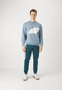 Felpa blu con design curvo bianco, abbinata a pantaloni joggers teal. Sneakers bianche completano l'outfit. Stile semplice e atletico.