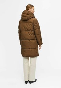 Doudoune marron avec capuche, dotée de sections matelassées horizontales et d'une coupe décontractée, assortie à un pantalon clair et des chaussures noires.