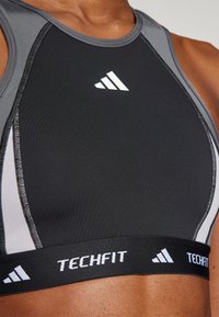 Svart og grå sportsbh med en strukturert design, glatt tekstur og elastisk midjebånd med "TECHFIT" merkevare og logoaksenter.