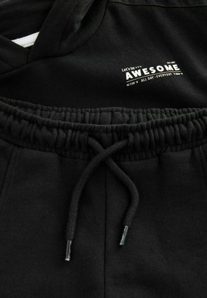 Hoodie noir avec un col à cordon, présentant un logo imprimé blanc. Tissu doux avec une coupe décontractée ; le pantalon a une taille élastique.