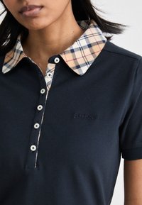 Tengerészkék pólóing, sötét kockás gallérral. Öt fehér gombbal rendelkezik, és a mellkasán sötét szálakkal hímzett "Barbour" logó található.