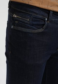 Jeans de mezclilla azul oscuro con un bolsillo frontal con cremallera, sutiles acentos de costura y un diseño de ajuste estándar.