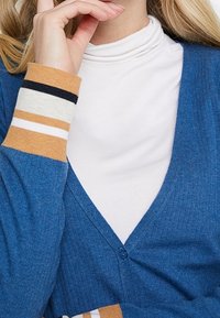 Blå ribbad kofta med V-hals och knappstängning. Har randiga detaljer i orange, marinblå och krämfärgad på ärmsluten. Vit topp med hög hals under.