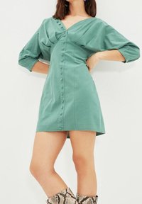 Vestido verde de manga corta con escote en V, pliegues horizontales y cierre de botones en la parte delantera. Presenta una silueta ajustada y una textura de tela suave.