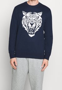 Sweat-shirt bleu marine avec un grand motif de tigre blanc accompagné du texte "SCHOTT FIGHTING SPIRIT" et du numéro "13". Mélange de coton, poignets et ourlet côtelés.