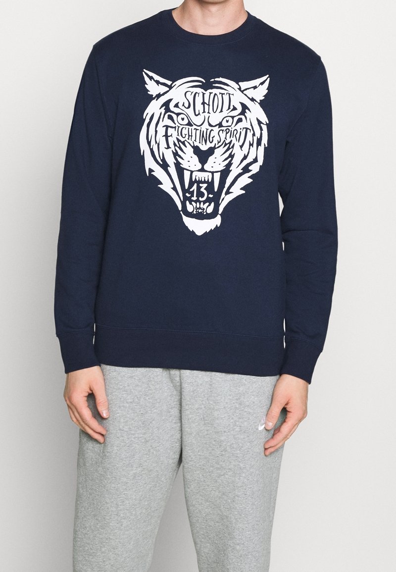 Sweat-shirt bleu marine avec un grand motif de tigre blanc accompagné du texte "SCHOTT FIGHTING SPIRIT" et du numéro "13". Mélange de coton, poignets et ourlet côtelés.