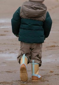 Child wearing a green and brown puffer jacket, beige cargo pants, and beige waterproof boots with blue accents, walking on a sandy beach.