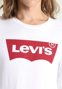 Weißes Baumwoll-T-Shirt mit langen Ärmeln, das ein großes rotes Levi's-Logo in einem markanten, geschwungenen Design über die Brustfläche zeigt.