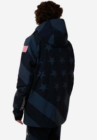 Giacca navy con cappuccio, caratterizzata da un motivo a stelle e strisce, tessuto resistente e marchio Kappa sulla manica. Dettaglio della toppa della bandiera americana.