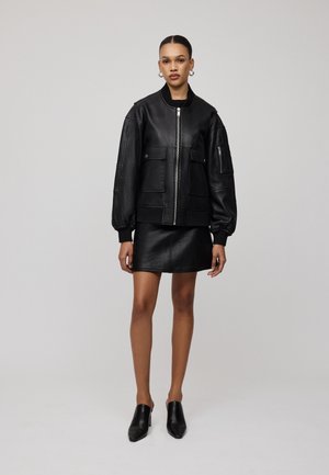 Giubbotto bomber in pelle nera con maniche oversize, tasche frontali e chiusura con zip, abbinato a una mini gonna in pelle coordinata e mules nere.