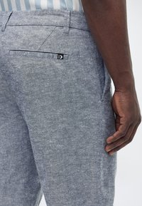 Εγγύς πλάνο ατόμου που φοράει ανοιχτό μπλε υφασμάτινο παντελόνι με πίσω τσέπη και ριγωτό πουκάμισο μέσα, με το χέρι ακουμπισμένο στο πλάι.