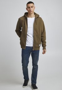 Khaki Zip-Hoodie mit Kapuze und Kordelzug, zwei Fronttaschen und einem weißen Rundhalsshirt darunter, kombiniert mit blauen Jeans und schwarzen Sneakers.