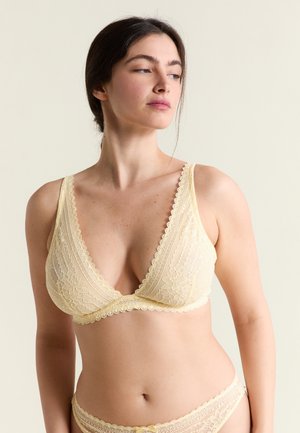 Mujer vistiendo un sostén de encaje amarillo pálido y ropa interior a juego, mirando hacia su izquierda sobre un fondo claro y uniforme.