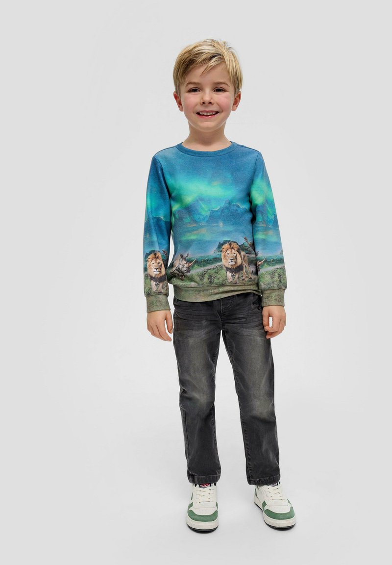 Blaues Langarm-Sweatshirt mit Löwengrafiken vor einer Landschaft mit Bergen, kombiniert mit dunkelgrauen Jeans und weiß-grünen Sneakers.