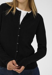 Svart ribbad kofta med knappar fram, långa ärmar och en mjuk textur. Bärs över en vit skjorta med minimalistiska designelement.